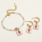 Seraphina Treasures Clytia bracelet and earring set with faux pearls, gold accents, and strawberry pendants on white squares.